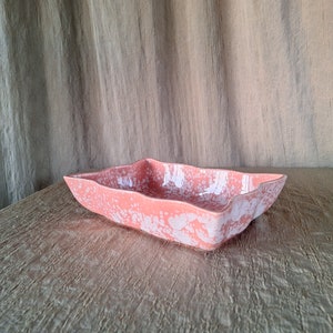 May include: A pink ceramic serving dish with a white speckled pattern. The dish has a wavy edge and is sitting on a textured beige surface.