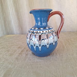 May include: A blue ceramic pitcher with a brown handle and white and brown painted designs. The pitcher has a round body and a narrow neck.