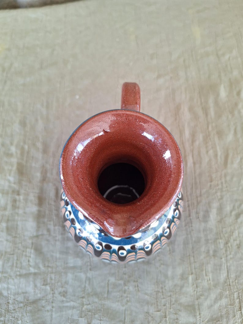 May include: A brown ceramic pitcher with a blue and white geometric pattern. The pitcher has a wide mouth and a handle.