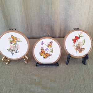 May include: Three embroidered hoop art pieces with butterfly designs. Each hoop is mounted on a small wooden stand. The butterflies are embroidered in shades of orange, yellow, brown, and red.