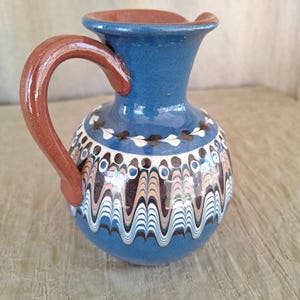 May include: A blue ceramic pitcher with a brown handle and white and brown painted designs. The pitcher has a rounded body and a narrow neck.