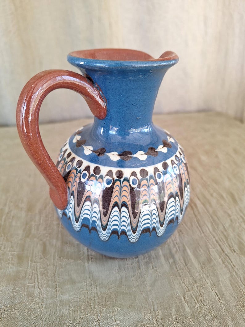 May include: A blue ceramic pitcher with a brown handle and white and brown painted designs. The pitcher has a rounded body and a narrow neck.