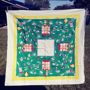 May include: A white tablecloth with a green and yellow border. The center of the tablecloth features a repeating pattern of red, yellow, and black images of kitchen items, including a stove, a teapot, a coffee pot, and a frying pan.