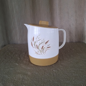 May include: White pitcher with a brown and green floral design. The pitcher has a lid and a handle. The pitcher is sitting on a brown textured surface.