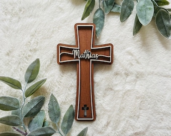 May include: A brown wooden cross featuring a layered design with a white border. The name Mathias is written in white cursive script across the horizontal bar, and a small cutout cross is positioned near the bottom of the vertical section.