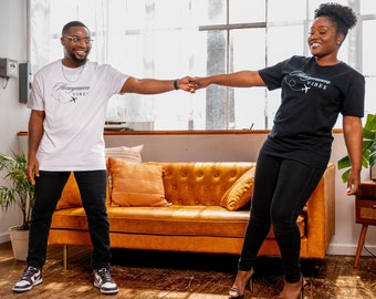 May include: A couple wearing matching black and white t-shirts with the text "Honeymoon Vibes" and a plane graphic. They are standing in a room with a brown sofa and wooden floors.