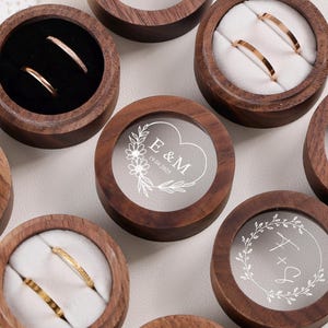 Könnte beinhalten: Eine Sammlung von Holz-Ringboxen mit transparenten Deckeln. Einige Boxen enthalten goldene Ringe. Die Deckel sind mit Initialen, Daten und Hochzeitsring-Grafiken graviert. Die Boxen sind rund.