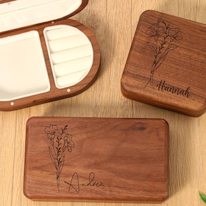 May include: Three wooden jewelry boxes. One is oval-shaped and open, revealing a white interior. Two are rectangular, one square, each with a floral design and a name engraved: "Hannah" and "Andrea".