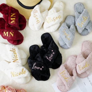 May include: A selection of plush cross-band slippers in burgundy, white, black, grey, and pink, personalised with names and initials in gold or pink script. A straw sun hat is in the background. A bouquet of pink roses is in the foreground.