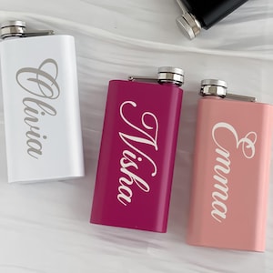 May include: A collection of personalized flasks in various colors. The flasks are rectangular with a silver cap and are engraved with names in a cursive font. Colors include white, black, pink, and magenta. The names visible are Olivia, Dani, Nisha, and Emma.