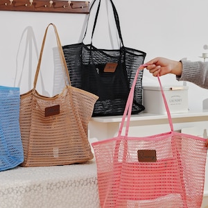 Könnte beinhalten: Vier Netztaschen in Blau, Beige, Schwarz und Pink. Jede Tasche hat einen rechteckigen Aufnäher und lange Henkel. Die blaue Tasche enthält ein Buch. Die Taschen stehen auf einer weißen Oberfläche.