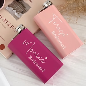 Könnte beinhalten: Eine Sammlung personalisierter Flachmänner in Rosa, Magenta und Weiß, jeweils mit silbernem Verschluss. Die Flachmänner sind mit Namen und dem Wort "Bridesmaid" beschriftet. Eine rosafarbene Cat-Eye-Sonnenbrille liegt auf einem Buch.