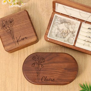 May include: Wooden jewellery boxes, one open, displaying necklaces and rings. Two closed boxes feature floral designs and names: "Kara" and "Claire." The boxes are a warm brown colour, with a light-coloured interior in the open box.
