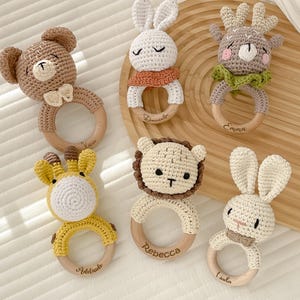 Könnte beinhalten: Sechs handgefertigte Babyrasseln aus Häkelarbeit in Tierform: ein Bär, zwei Hasen, ein Reh, eine Giraffe und ein Löwe. Jede Rassel hat einen Holzring und ist in neutralen Farben gehalten.