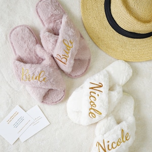 May include: A pair of light pink fluffy slippers embroidered with the word "Bride" in gold, and a pair of white fluffy slippers embroidered with the name "Nicole" in gold. A straw hat with a black band is also in the image.