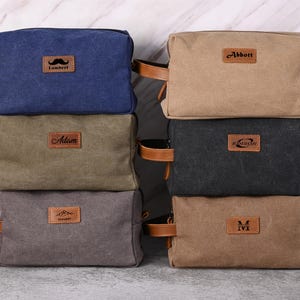 May include: A stack of six canvas toiletry bags in various colors: navy blue, khaki, olive green, charcoal gray, and brown. Each bag has a leather handle and a small leather patch with a name or initial. The bags are rectangular and have a zipper closure.