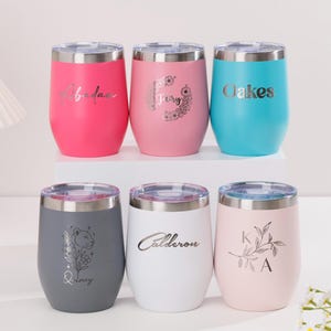 Op de afbeelding: Zes geïsoleerde, steelvrije wijnglazen in verschillende kleuren: roze, lichtroze, blauw, grijs en wit. Elke beker heeft een zilveren deksel en is voorzien van gepersonaliseerde tekst of een bloemmotief. De bekers zijn ontworpen om dranken koud te houden.