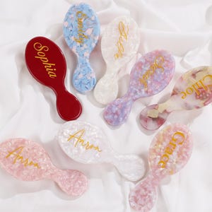May include: An assortment of personalized hairbrushes in various colors, including red, blue, pink, and white. Each brush features a unique name in gold script. The brushes have an oval head and a handle.