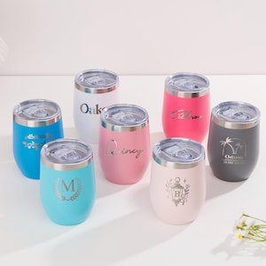 Può includere: Una collezione di bicchieri da vino isolati in vari colori, tra cui blu, rosa, bianco e grigio. Ogni bicchiere ha un coperchio argentato e presenta testo o disegni personalizzati. I bicchieri sono disposti su una superficie bianca.