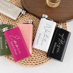 Peut inclure: Plusieurs flasques personnalisées de couleurs variées : argent, vert olive, rose, fuchsia, blanc et noir. Chaque flasque porte un nom et le mot "Bridesmaid" ou "Maid of Honor". Un petit flacon de parfum est posé sur un plateau en bois.