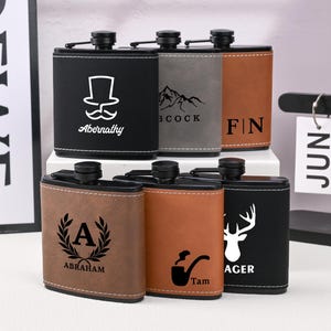 Può includere: Una collezione di fiaschette personalizzate in vari colori, tra cui nero, marrone e grigio. Ogni fiaschetta presenta disegni e testi unici, come un cappello a cilindro e baffi, montagne, iniziali e una testa di cervo. Le fiaschette sono realizzate in materiale simile alla pelle.
