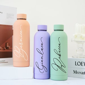 May include: Three personalized water bottles with names "Bernice", "Gianluca", and "Rebecca" written in cursive. The bottles are peach, lavender, and green.