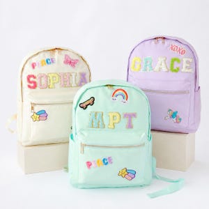 May include: Three colorful backpacks with embroidered patches. One is cream-colored with the name "SOPHIA" and the word "PEACE". Another is mint green with "MPT" and "PEACE". The third is lavender with "GRACE" and "XOXO". Each has a front pocket.