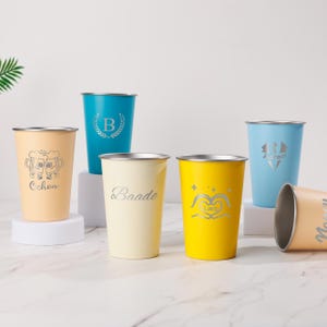Può includere: Una collezione di bicchieri in acciaio inossidabile in vari colori, tra cui pesca, turchese, crema e giallo. Ogni bicchiere presenta un bordo argentato e incisioni personalizzate, come nomi, iniziali e disegni grafici. I bicchieri sono esposti su una superficie bianca.