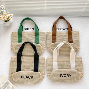 May include: Four straw tote bags with different colored handles: green, brown, black, and ivory. Each bag has a rectangular shape and the name of the handle color printed on the front. The bags are made of woven straw material.