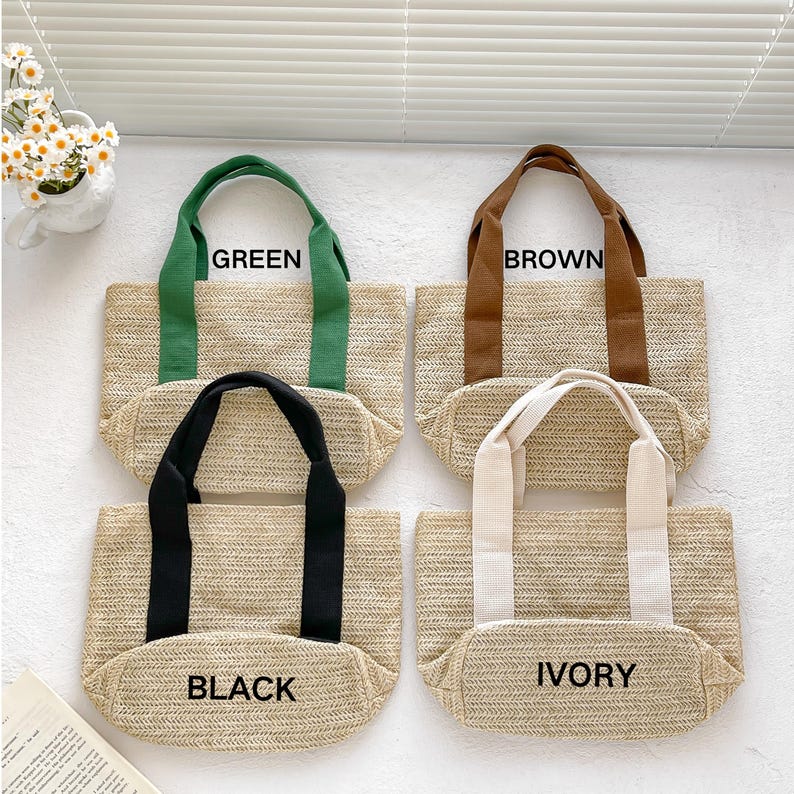 May include: Four straw tote bags with different colored handles: green, brown, black, and ivory. Each bag has a rectangular shape and the name of the handle color printed on the front. The bags are made of woven straw material.