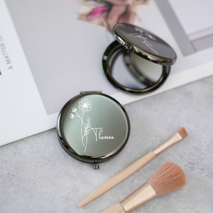 Op de afbeelding: Een zwarte compact spiegel met een bloemenontwerp en de naam "Theresa" gegraveerd op de voorkant. De spiegel is open en toont een reflectie. Twee make-upkwasten zijn ook inbegrepen.