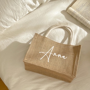 May include: A rectangular jute tote bag with the name "Anna" in white script. The bag has cream-colored handles and is sitting on a white surface. A book is visible in the background.