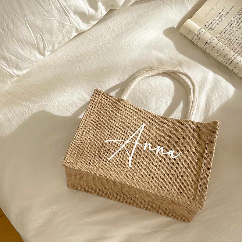 May include: A rectangular jute tote bag with the name "Anna" in white script. The bag has cream-colored handles and is sitting on a white surface. A book is visible in the background.