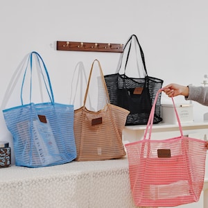 Puede incluir: Cuatro bolsas de malla en azul, beige, negro y rosa. Cada bolsa tiene un parche rectangular y asas largas. La bolsa azul contiene un libro. Las bolsas est&aacute;n sobre una superficie blanca.