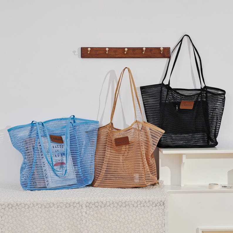Puede incluir: Tres bolsos de mano de malla en azul, beige y negro. Cada bolso tiene un parche rectangular de cuero y asas largas. Los bolsos se exhiben sobre una superficie blanca contra una pared blanca, con un perchero de madera con ganchos en el fondo.