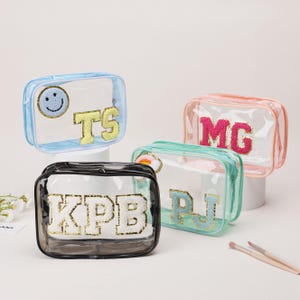 May include: Four clear cosmetic bags with embroidered letters and designs. The bags are in various colours: blue, pink, green, and black. Each bag has a different initial or design, such as a smiley face or initials.