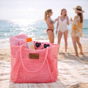 Puede incluir: Una bolsa de playa de malla rosa llena de una toalla rosa, protector solar y gafas de sol en forma de coraz&oacute;n. La bolsa tiene una etiqueta de cuero marr&oacute;n con la palabra "Fairy". La bolsa est&aacute; sobre una toalla de playa en la arena, con el mar y tres personas al fondo.