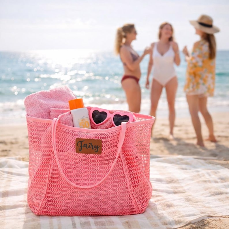 Puede incluir: Una bolsa de playa de malla rosa llena de una toalla rosa, protector solar y gafas de sol en forma de coraz&oacute;n. La bolsa tiene una etiqueta de cuero marr&oacute;n con la palabra "Fairy". La bolsa est&aacute; sobre una toalla de playa en la arena, con el mar y tres personas al fondo.