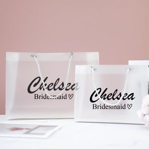 May include: Two clear plastic gift bags with white handles. The bags have the name "Chelsea" and the word "Bridesmaid" printed on them.