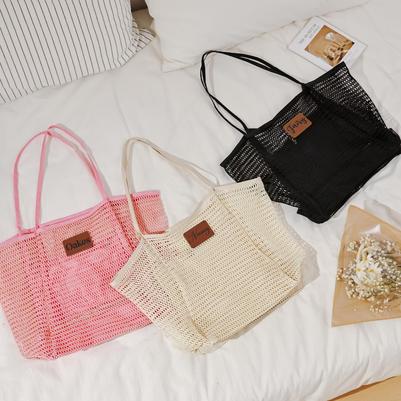 Puede incluir: Tres bolsas de malla en rosa, crema y negro, cada una con una etiqueta rectangular de polipiel. La bolsa rosa tiene "Oakes" en la etiqueta, la bolsa crema "Nancy" y la bolsa negra "Fairy". Las bolsas tienen asas largas.