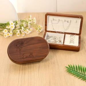 May include: A wooden jewelry box with an open lid, revealing compartments for rings and necklaces. The box is made of dark wood and has a floral design and the name "Emilh" engraved on the lid. The interior is lined with white fabric.