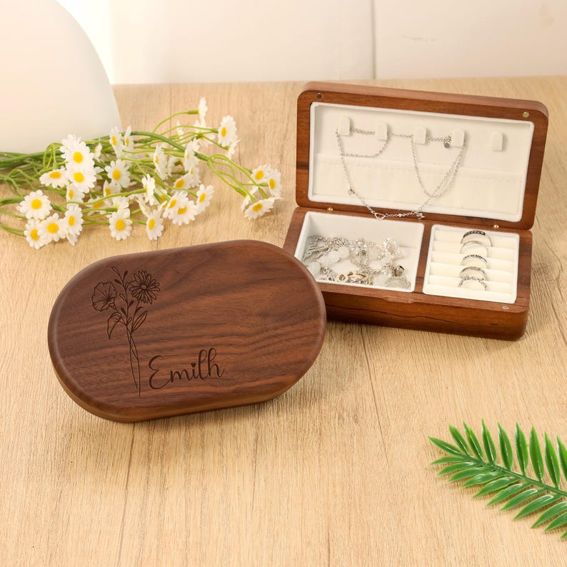 May include: A wooden jewelry box with an open lid, revealing compartments for rings and necklaces. The box is made of dark wood and has a floral design and the name "Emilh" engraved on the lid. The interior is lined with white fabric.