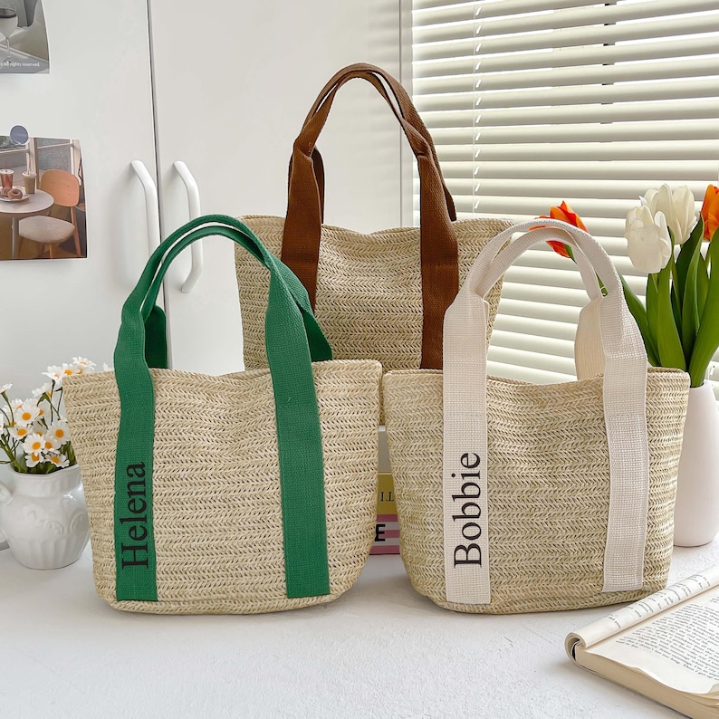 May include: Three woven straw tote bags with different colored handles. The bag on the left has green handles and the name "Helena" printed on the front. The middle bag has brown handles. The bag on the right has white handles and the name "Bobbie" printed on the front.
