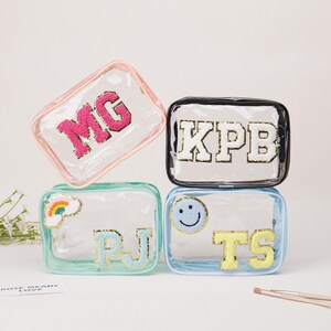 May include: Four clear cosmetic bags with different colored trim. Each bag has a different embroidered design: "MG" in pink, "KPB" in white, "PJ" in light blue, and "TS" in yellow. A rainbow and a smiley face are also embroidered on two of the bags.