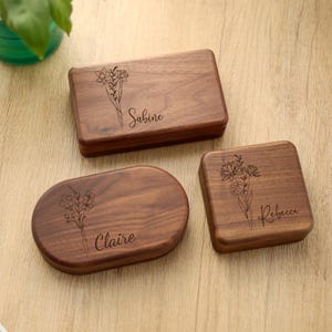 May include: Three wooden boxes with floral engravings and names: "Sabine", "Claire", and "Rebecca". The boxes are in different shapes: rectangular, oval, and square. The wood has a warm, brown tone.