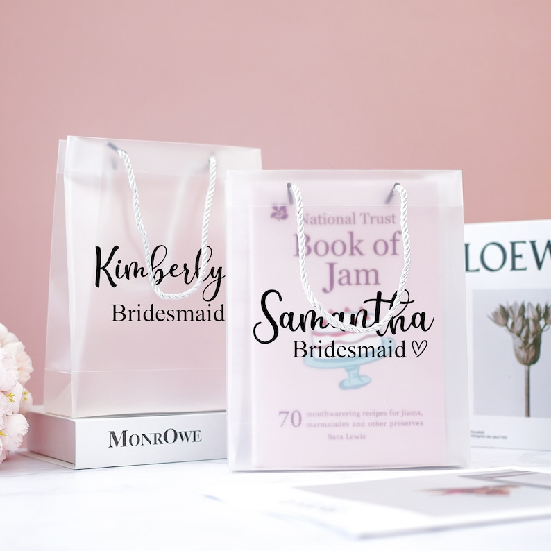 May include: Two clear plastic gift bags with white handles. The bags are decorated with black text that says "Kimberly Bridesmaid" and "Samantha Bridesmaid♡". The bag on the right also has a book inside with the title "National Trust Book of Jam".