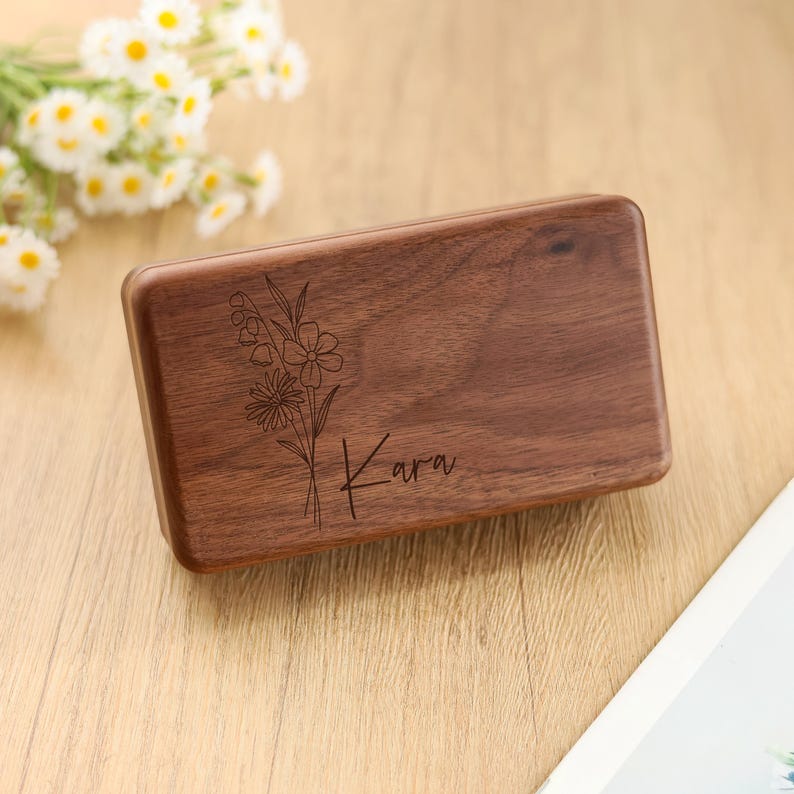 May include: A rectangular wooden box with a floral design and the name "Kara" engraved on the surface. The box is made of dark brown wood and has a smooth finish. The floral design includes various flower types.