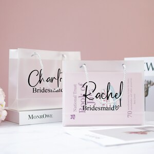 May include: Two clear plastic gift bags with white handles. The bags are filled with gifts and have personalized labels that say "Charlotte Bridesmaid" and "Rachel Bridesmaid".