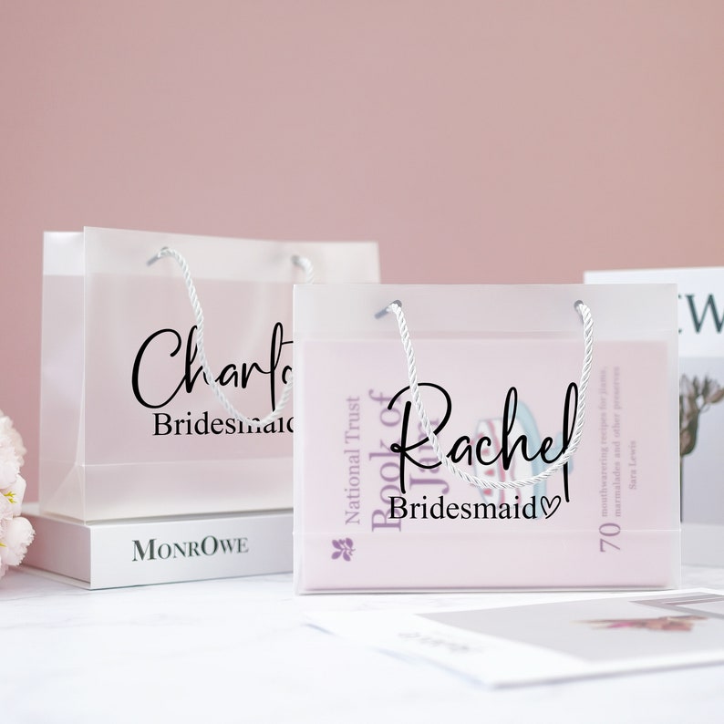 May include: Two clear plastic gift bags with white handles. The bags are filled with gifts and have personalized labels that say "Charlotte Bridesmaid" and "Rachel Bridesmaid".