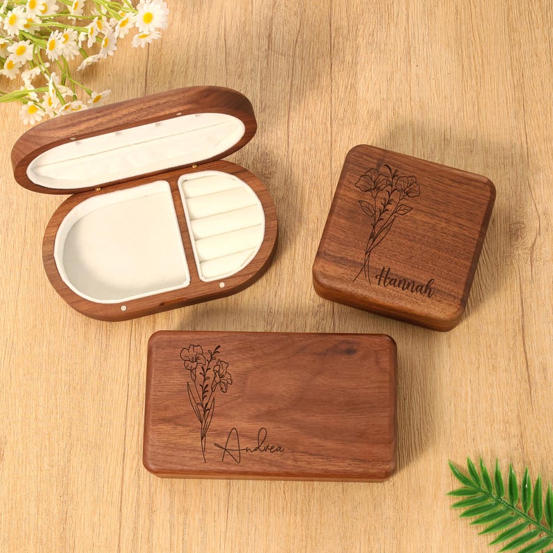 May include: Three wooden jewelry boxes. One is oval-shaped and open, revealing a white interior. Two are rectangular, one square, each with a floral design and a name engraved: "Hannah" and "Andrea".